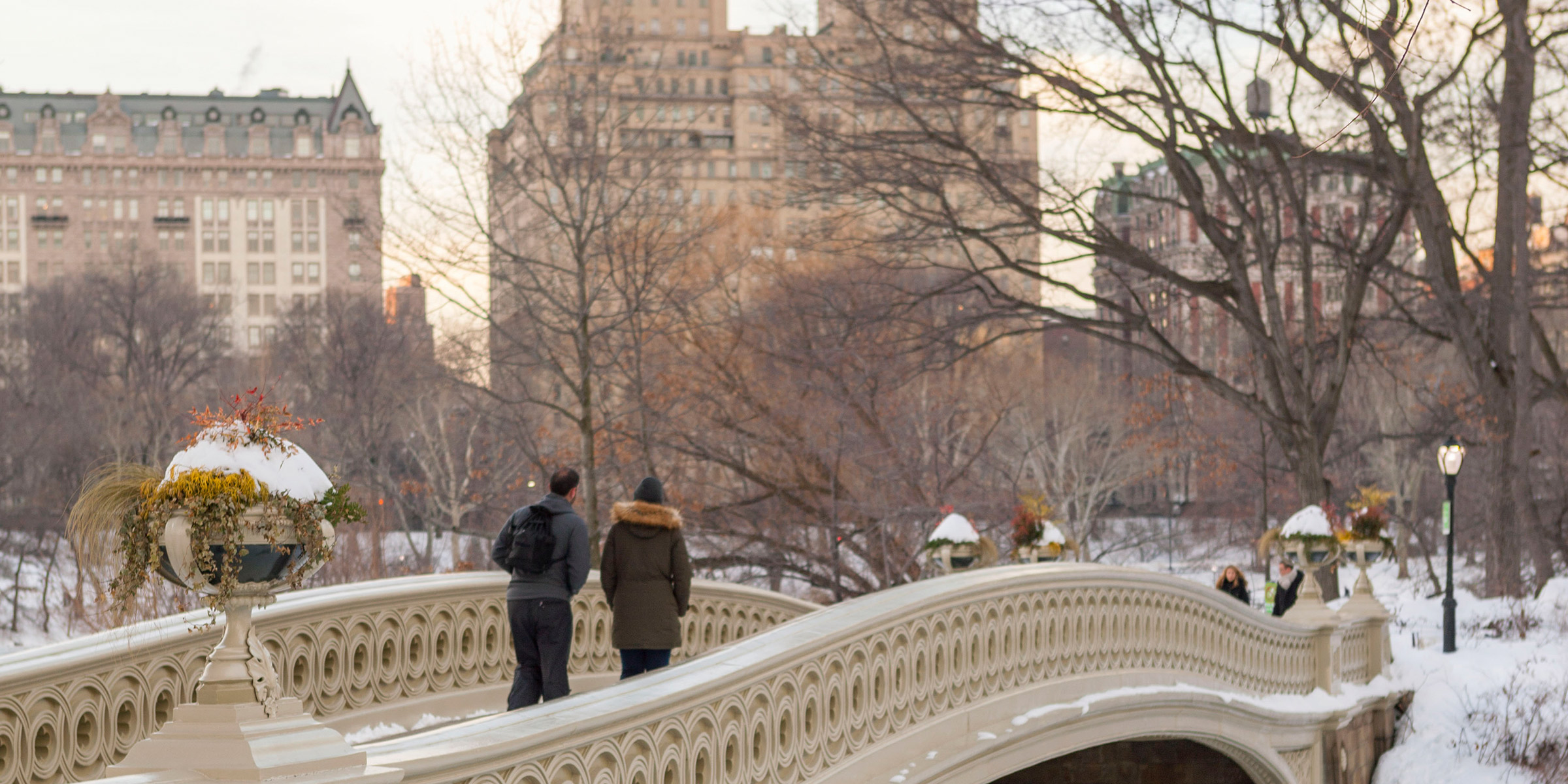 Snowy Bow Bridge in Central Park with city backdrop.