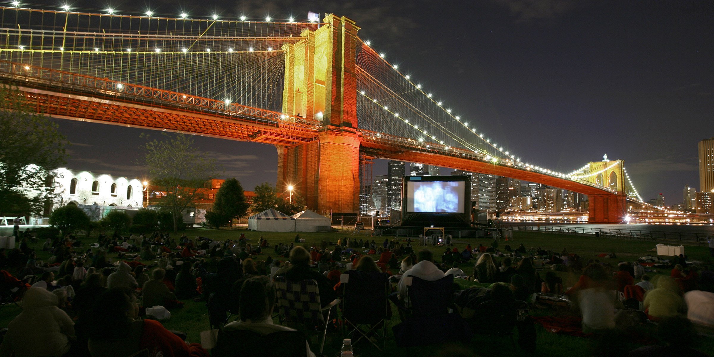 Outdoor movie night under Brooklyn Bridge with city lights.