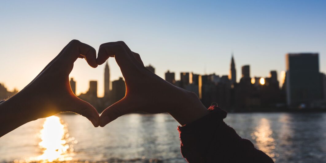 Hands forming a heart shape against the New York City skyline at sunset.