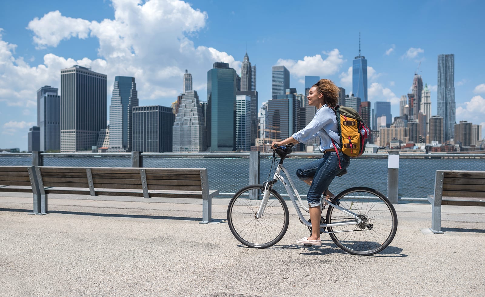 Woman riding a bicycle along a waterfront with the New York City skyline.