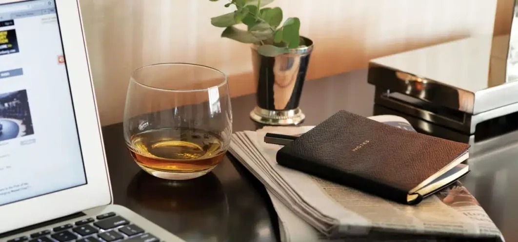 Whiskey glass, notebook, and laptop on desk.